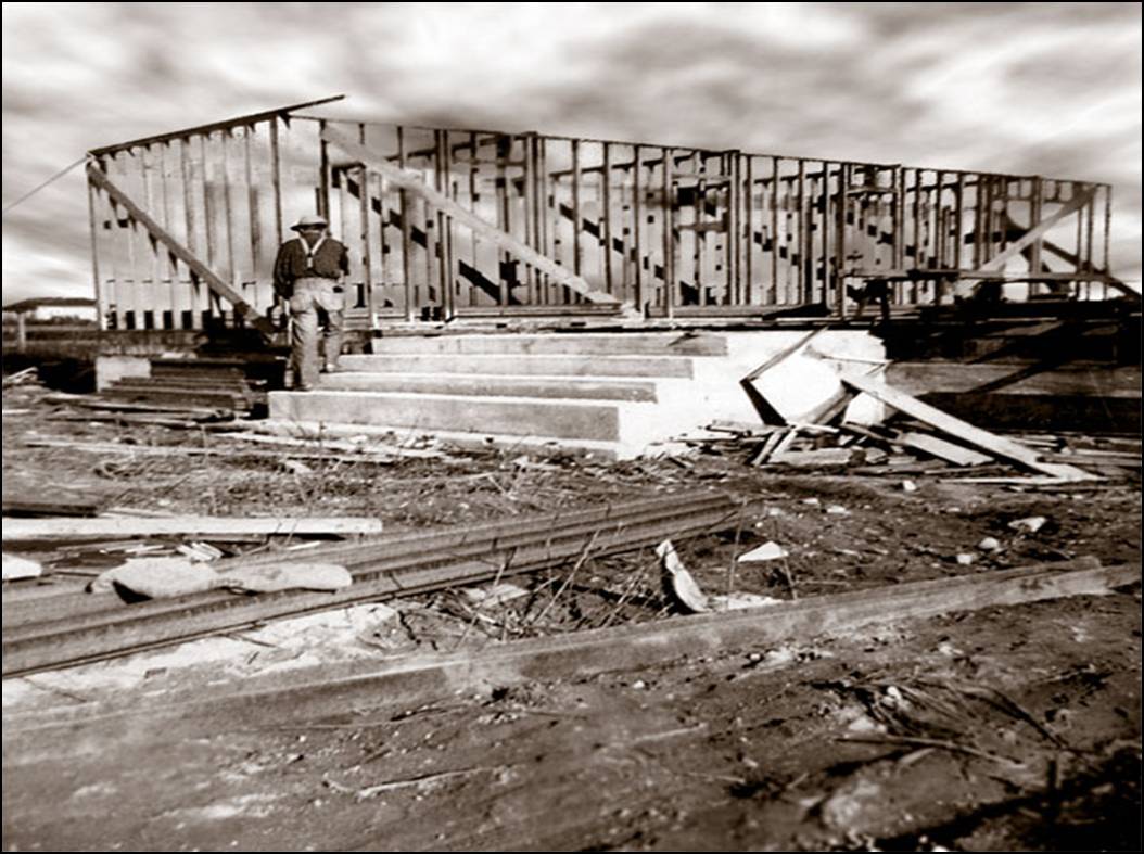 Historic 1955 black and white photograph showing the structural framing stage of the parish hall, with a construction worker performing framing work on a primary exterior wall.