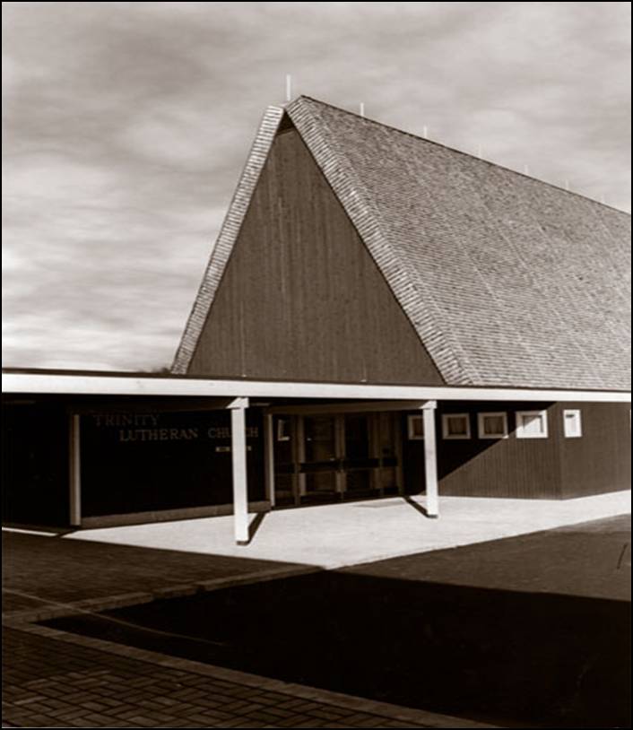 A 1964 historic photograph looking northwest at the newly completed Trinity Lutheran Church in Richmond, BC. The flat-roofed entrance and office wing with vertical wood siding are in the foreground, while the massive shingled south elevation of the A-frame sanctuary rises behind it. Modern paving and a covered walkway lead to the main doors.